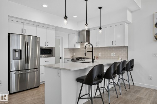 Bright kitchen featuring a central island with countertop seating, white cabinetry, stainless steel appliances, a light-toned tile backsplash, and wood-finish flooring - 23027 96 Avenue Nw, Edmonton, AB - Indoor Photo Showing Kitchen With Stainless Steel Kitchen With Upgraded Kitchen