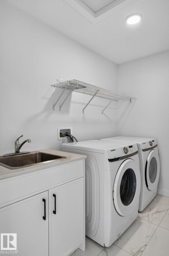 Dedicated laundry room featuring white walls, a white wire shelving unit, a stainless steel utility sink, white base cabinetry with black hardware, and marble-look floor tile - 23027 96 Avenue Nw, Edmonton, AB - Indoor Photo Showing Laundry Room