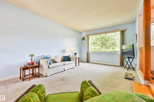 Spacious living area featuring light blue walls, a large picture window, light-toned carpeting, and a wood banister with vertical spindles - 3824 111B Street Nw, Edmonton, AB - Indoor