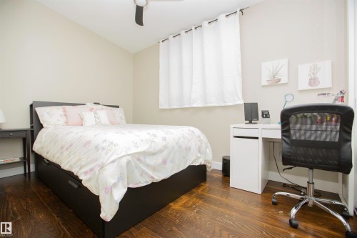 Bright bedroom featuring a wood-finish floor, black platform bed frame, large window with sheer curtains allowing natural light, white built-in desk with integrated storage, and mesh office chair with chrome base - 9107 58 Street Nw, Edmonton, AB - Indoor Photo Showing Bedroom