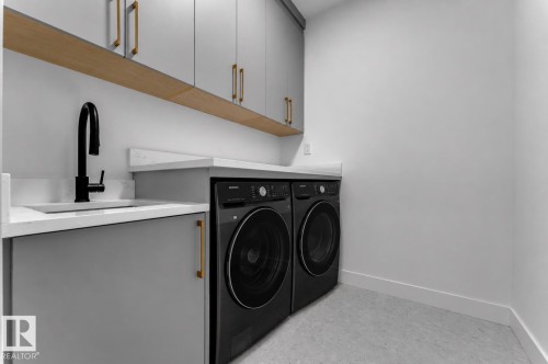 Laundry area featuring a built-in sink with a matte black faucet, light-toned countertops, grey cabinetry with brass hardware, and integrated appliances - 2039 156 Avenue Nw, Edmonton, AB - Indoor Photo Showing Laundry Room