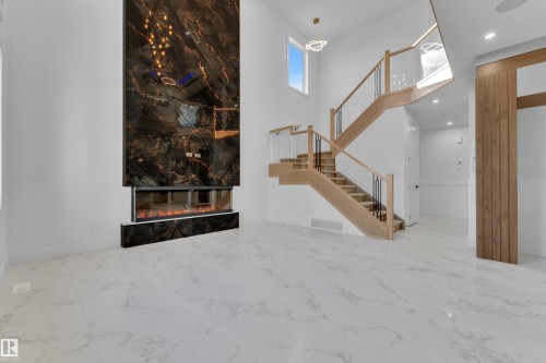 Sleek linear fireplace with dark marble-finish surround, light wood-finish staircase with glass railings, white marble-look flooring, and a contemporary ceiling light fixture - 2039 156 Avenue Nw, Edmonton, AB - Indoor Photo Showing Other Room