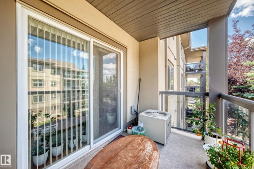 Private balcony featuring a sliding glass door with vertical blinds, a glass and metal railing, and an exterior wall-mounted air conditioning unit - #324 279 Suder Greens Dr Nw, Edmonton, AB - Outdoor With Balcony With Exterior
