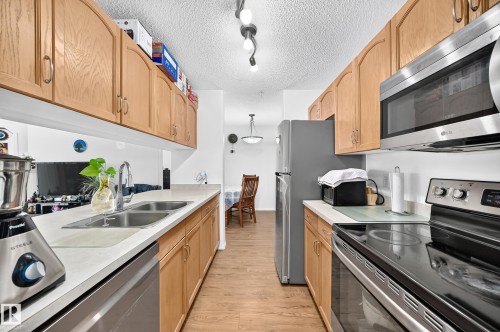 Galley kitchen featuring wood-finish cabinetry, stainless steel appliances, light countertops, and wood-finish flooring - #324 279 Suder Greens Dr Nw, Edmonton, AB - Indoor Photo Showing Kitchen With Double Sink