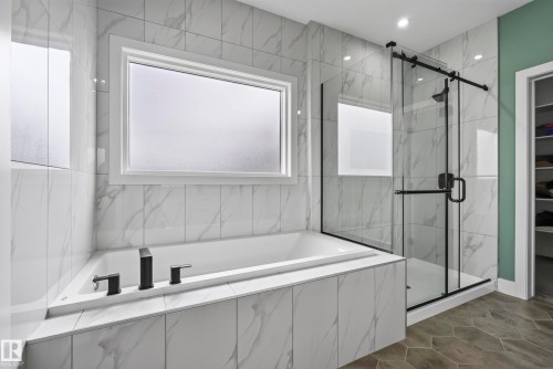Soaking tub with matte black fixtures, an adjacent glass-enclosed shower with a black frame, white tiled walls with subtle veining, and hexagonal floor tiling - 7314 Morgan Road Nw, Edmonton, AB - Indoor Photo Showing Bathroom
