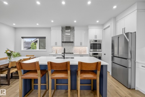 Kitchen featuring a central island with countertop seating, stainless steel appliances, white cabinetry, and wood-finish flooring - 7314 Morgan Road Nw, Edmonton, AB - Indoor