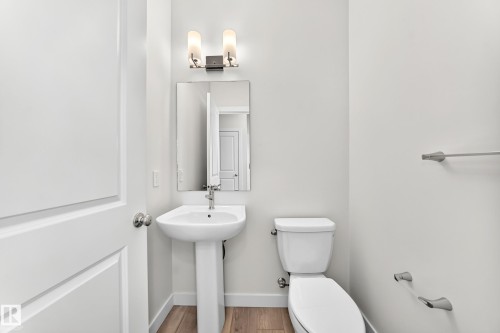 Pedestal sink with chrome faucet, wall-mounted mirror, two-light vanity fixture, white toilet, and wood-finish flooring - 3816 Chrustawka Place Sw, Edmonton, AB - Indoor Photo Showing Bathroom