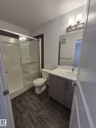Enclosed shower with sliding glass doors, white toilet, vanity with integrated sink, wood-finish flooring, and wall-mounted light fixture - Edmonton, AB - Indoor Photo Showing Bathroom