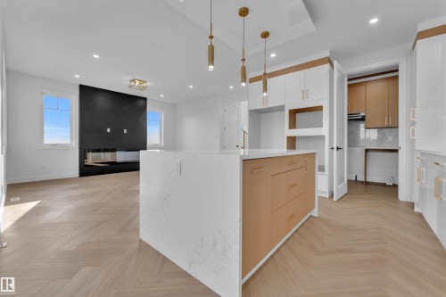 Open-concept living space with herringbone wood-finish flooring, a kitchen island featuring a light-toned wood finish and a white countertop, and a modern fireplace with a dark surround - 465 Crystal Creek Link, Leduc, AB - Indoor Photo Showing Other Room