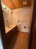 Master bathroom featuring dual vanity with dark cabinetry and countertop, framed mirror with integrated lighting, ceramic tile shower enclosure with glass partition and chrome fixtures, and herringbone wood-finish flooring throughout - 