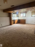 Spacious basement recreation area featuring exposed wood-beam ceiling, carpeted flooring, built-in cabinetry with wood finishes, and multiple windows providing natural light - 