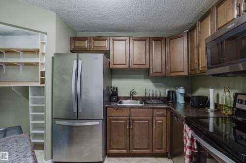 Stainless steel appliances, wood-finish cabinetry, built-in shelving, corner sink with faucet, and dark countertops - #1002 10149 Saskatchewan Dr Nw, Edmonton, AB - Indoor Photo Showing Kitchen