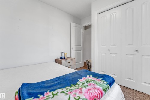 White bi-fold closet doors with panel detailing, light wood-finish nightstand with two drawers, neutral-toned carpeting, and a white interior door - 2040 24 Street Nw, Edmonton, AB - Indoor Photo Showing Bedroom