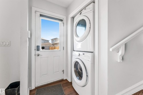 White stacked washer and dryer unit, wood-finish flooring, white door with upper window, white wall-mounted handrail, and light grey wall paint - 2040 24 Street Nw, Edmonton, AB - Indoor Photo Showing Laundry Room