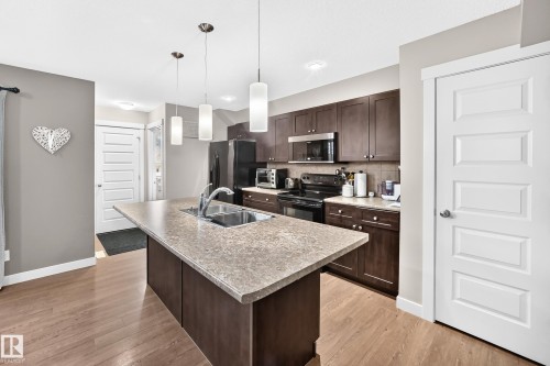 Kitchen featuring a central island with an undermount double basin sink, dark wood-finish cabinetry, black appliances, light-toned countertops, and wood-finish flooring - 466 Chappelle Drive Sw, Edmonton, AB - Indoor Photo Showing Kitchen With Double Sink With Upgraded Kitchen