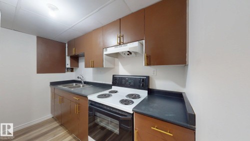 Kitchen featuring wood-finish cabinetry with golden hardware, a double basin stainless steel sink, dark countertops, a white electric range, and wood-finish flooring - 2426 33A Street Nw, Edmonton, AB - Indoor Photo Showing Kitchen With Double Sink