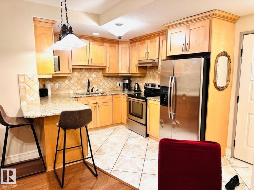 Light wood cabinetry with crown molding, granite-style countertops, stainless steel appliances, a tiled backsplash, and tile flooring - #207 10142 111 St Nw, Edmonton, AB - Indoor Photo Showing Kitchen