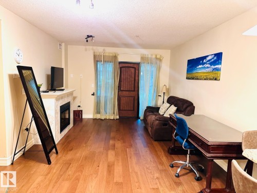 Wood-finish flooring, a fireplace with a light-toned tile surround and mantel, and a dark wood entry door with vertical panels - #207 10142 111 St Nw, Edmonton, AB - Indoor Photo Showing Living Room With Fireplace