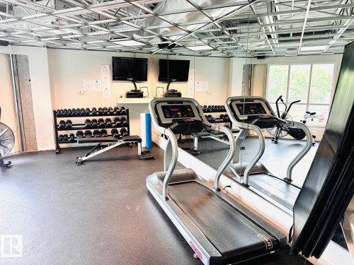Fitness center with rubber flooring, exposed ceiling grid, and multiple wall-mounted televisions - #207 10142 111 St Nw, Edmonton, AB - Indoor Photo Showing Gym Room