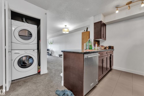 Full-size stacked laundry appliances, dark wood-finish cabinetry, stainless steel dishwasher, and track lighting - #409 11804 22 Av Sw, Edmonton, AB - Indoor Photo Showing Laundry Room