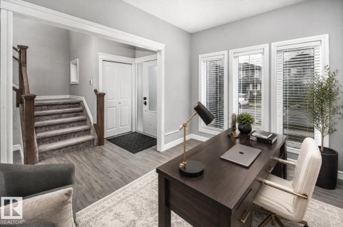 Wood-finish flooring throughout, bright entryway with a paneled door and sidelight, staircase with wood newel posts and carpeted treads, and an office space featuring three large windows with blinds - 16015 11 Avenue Sw, Edmonton, AB - Indoor Photo Showing Other Room