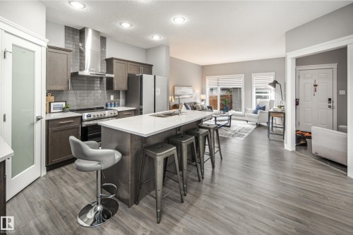 Kitchen island with a white countertop and seating, stainless steel appliances, gray subway tile backsplash, recessed lighting, and wood-finish flooring - 16015 11 Avenue Sw, Edmonton, AB - Indoor Photo Showing Kitchen With Upgraded Kitchen
