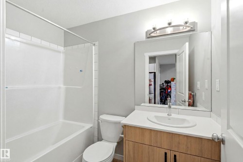 Bathroom featuring a white tub/shower combination, a vanity with wood-finish cabinetry, a white countertop, and a rectangular undermount sink with a chrome faucet - #106 856 Secord Bv Nw Nw, Edmonton, AB - Indoor Photo Showing Bathroom