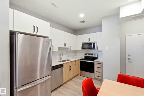 Kitchen featuring stainless steel appliances, white upper cabinetry, wood-finish lower cabinetry, light-toned countertops, and subway tile backsplash - #106 856 Secord Bv Nw Nw, Edmonton, AB - Indoor Photo Showing Kitchen With Double Sink