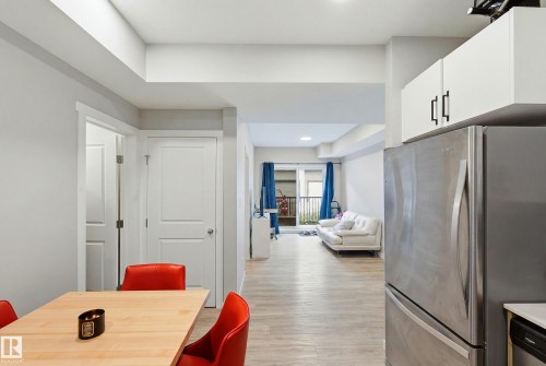 Integrated kitchen with stainless steel refrigerator, white upper cabinetry, and wood-finish flooring extending into the living area - #106 856 Secord Bv Nw Nw, Edmonton, AB - Indoor
