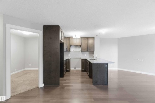 Open-concept kitchen featuring dark wood cabinetry, light-toned countertops, an island with a sink, and integrated appliances - #114 2207 44 Av Nw, Edmonton, AB - Indoor Photo Showing Kitchen