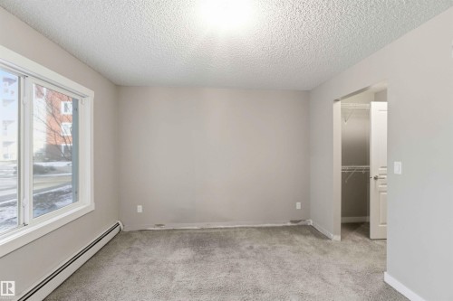 Carpeted room featuring a large window with white trim, baseboard heating, a neutral wall color, and an open doorway revealing a closet with wire shelving - #114 2207 44 Av Nw, Edmonton, AB - Indoor Photo Showing Other Room
