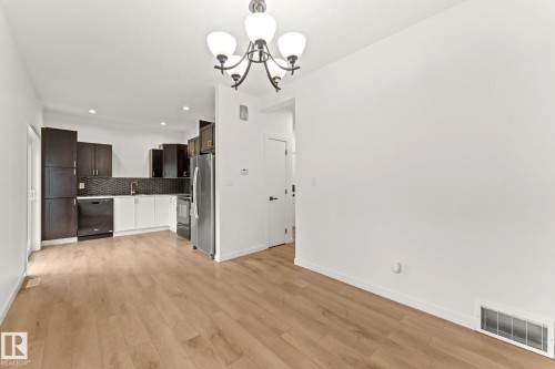 Open-concept living area with wood-finish flooring, modern kitchen featuring dark upper cabinetry and white lower cabinetry, black tile backsplash, stainless steel appliances, and a contemporary chandelier - 10825 93 Street Nw, Edmonton, AB - Indoor Photo Showing Kitchen