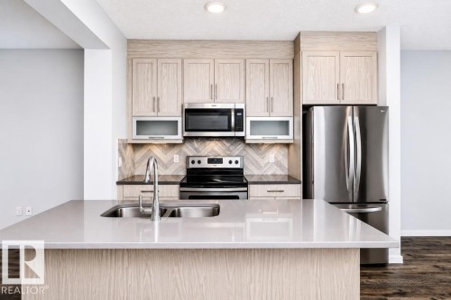Kitchen island with a double basin stainless steel sink, wood-finish cabinetry, stainless steel appliances, and a tiled backsplash - #6 215 Saddleback Rd Nw, Edmonton, AB - Indoor Photo Showing Kitchen With Double Sink With Upgraded Kitchen