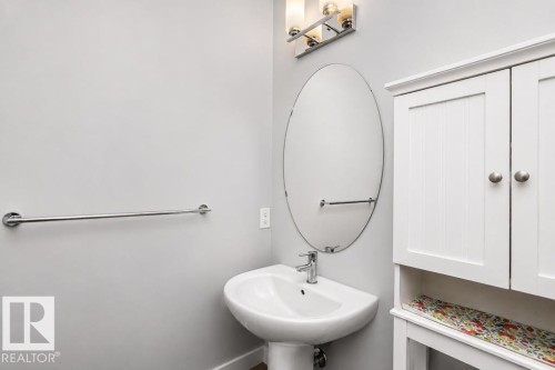 Pedestal sink with chrome faucet, oval mirror, wall-mounted light fixture, white storage cabinet with beadboard doors, and chrome towel bar - #6 215 Saddleback Rd Nw, Edmonton, AB - Indoor Photo Showing Bathroom