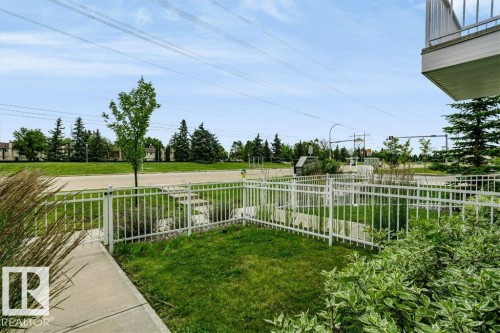 White metal fencing enclosing a landscaped green space with mature plantings and a concrete walkway - #6 215 Saddleback Rd Nw, Edmonton, AB - Outdoor