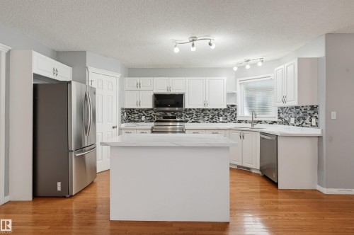 Modern kitchen featuring white cabinetry, stainless steel appliances, a large center island with white countertops, decorative tile backsplash, warm wood-finish flooring, and recessed and track lighting throughout - #8 1920 Millwoods Rd E Nw, Edmonton, AB - Indoor Photo Showing Kitchen With Upgraded Kitchen