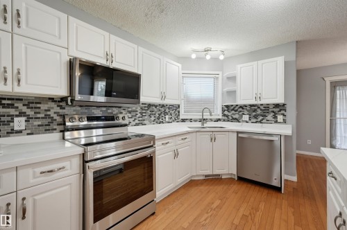 White cabinetry, stainless steel appliances, mosaic tile backsplash, solid surface countertops, and wood-finish flooring - #8 1920 Millwoods Rd E Nw, Edmonton, AB - Indoor Photo Showing Kitchen With Upgraded Kitchen