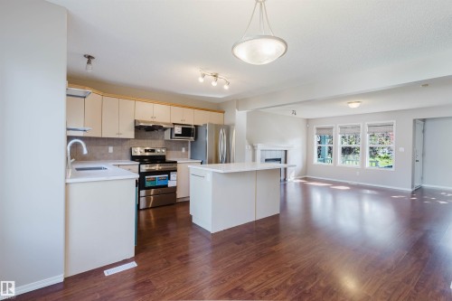 Wood-finish flooring throughout, kitchen with stainless steel appliances, central island with solid surface countertop, white cabinetry, and a fireplace with a mantel - 5887 Sutter Place Nw, Edmonton, AB - Indoor Photo Showing Kitchen With Stainless Steel Kitchen