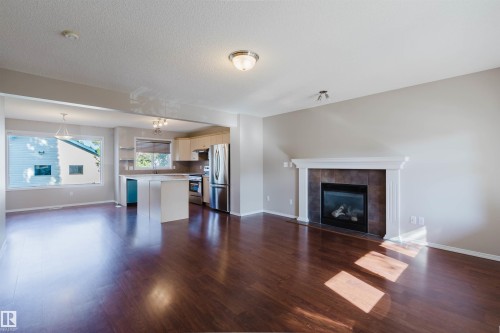Open-concept living area with dark wood-finish flooring, a white mantel fireplace with tile surround, and a kitchen featuring stainless steel appliances and light cabinetry - 5887 Sutter Place Nw, Edmonton, AB - Indoor Photo Showing Living Room With Fireplace