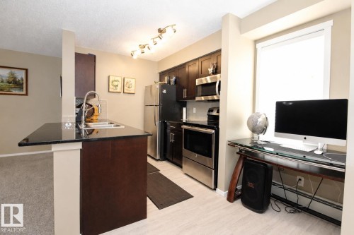 Kitchen featuring dark wood-finish cabinetry, stainless steel appliances, track lighting, and a black countertop island with an undermount sink - #236 3315 James Mowatt Tr Sw, Edmonton, AB - Indoor Photo Showing Kitchen With Double Sink