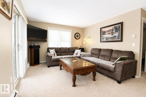 Wall-to-wall carpeting throughout, neutral painted walls, sliding glass door, and a built-in window with blinds - #236 3315 James Mowatt Tr Sw, Edmonton, AB - Indoor Photo Showing Living Room
