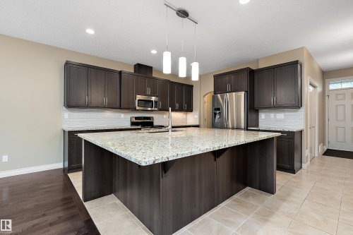 Spacious kitchen featuring a large island with a light-toned countertop, dark wood-finish cabinetry, stainless steel appliances, a white subway tile backsplash, and a modern pendant light fixture - 11203 72 Avenue Nw, Edmonton, AB - Indoor Photo Showing Kitchen With Upgraded Kitchen