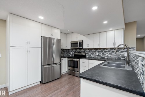Kitchen featuring white cabinetry, stainless steel appliances, dark countertops, a mosaic tile backsplash, and wood-finish flooring - 11203 72 Avenue Nw, Edmonton, AB - Indoor Photo Showing Kitchen With Double Sink With Upgraded Kitchen