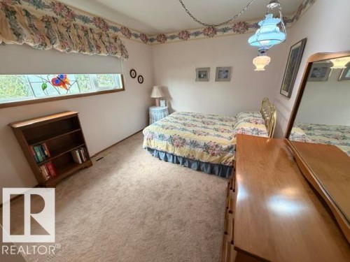 Carpeted interior space featuring a horizontal window with stained-glass details, a brass-tone bed frame, and a wood-finish dresser with an attached mirror - 6824 89 Avenue Nw, Edmonton, AB - Indoor Photo Showing Bedroom