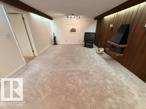 Expansive carpeted room featuring a brick fireplace with a black metal hood and wood paneling - 6824 89 Avenue Nw, Edmonton, AB - Indoor Photo Showing Other Room