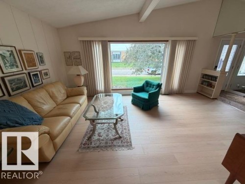 Vaulted ceiling with exposed beam, large picture window with vertical blinds, light wood-finish flooring, built-in shelving, and a decorative glass-top table - 6824 89 Avenue Nw, Edmonton, AB - Indoor Photo Showing Living Room