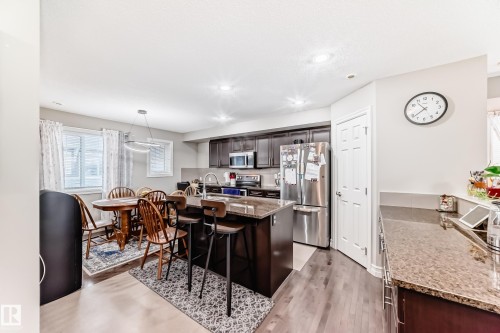 Open-concept kitchen featuring dark wood cabinetry, stainless steel appliances, a center island with bar seating, and stone-finish countertops - #138 655 Tamarack Rd Nw, Edmonton, AB - Indoor