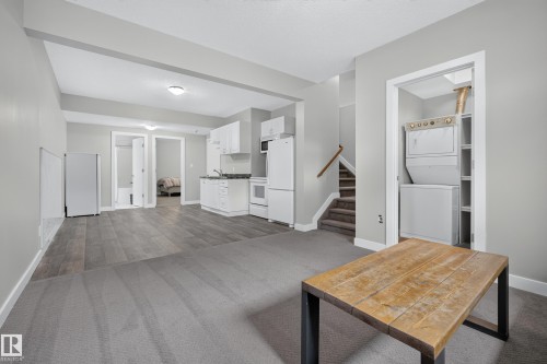 Spacious room featuring gray carpet and wood-finish flooring, a functional kitchen area with white cabinetry, a stackable washer and dryer unit, and a staircase with wood railing - 11018 122 Street Nw, Edmonton, AB - Indoor Photo Showing Other Room