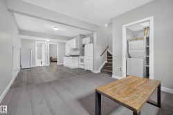 Spacious room featuring gray carpet and wood-finish flooring, a functional kitchen area with white cabinetry, a stackable washer and dryer unit, and a staircase with wood railing - 
