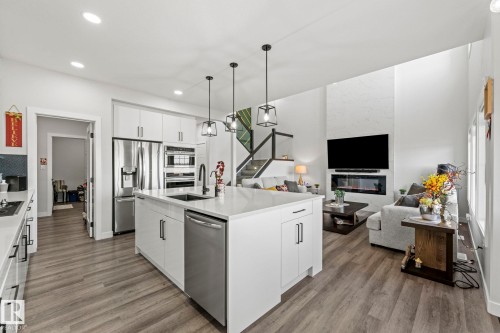 Open-concept living space with wood-finish flooring, white cabinetry, stainless steel appliances, and a built-in fireplace - 16128 31 Avenue Sw, Edmonton, AB - Indoor Photo Showing Kitchen With Upgraded Kitchen